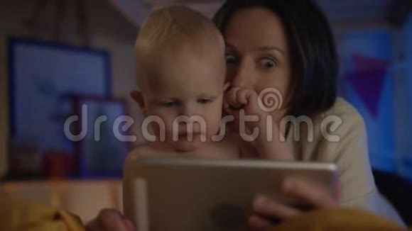 母亲在她的平板上看书给婴儿躺在床上上面画着不同的图画并给他糖果视频的预览图