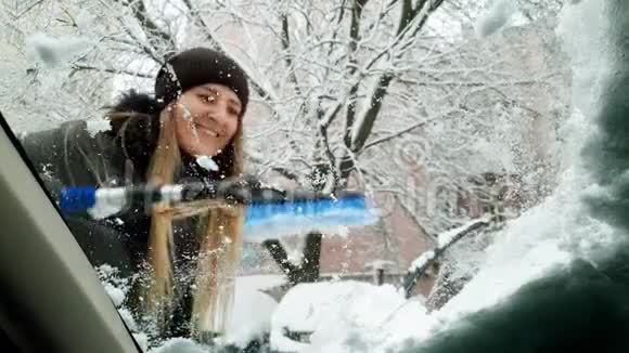 4k视角视频美丽的微笑年轻女子用刷子从雪中清洁汽车挡风玻璃视频的预览图
