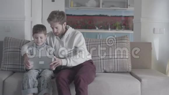大客厅里长胡子的父亲和孩子用数码平板电脑坐在沙发上爸爸教他的儿子父子关系视频的预览图