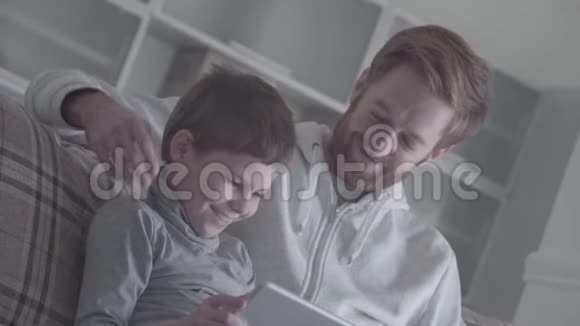 父亲和儿子一边使用数字小工具一边纵容地坐在沙发上爸爸和孩子之间的友好关系视频的预览图