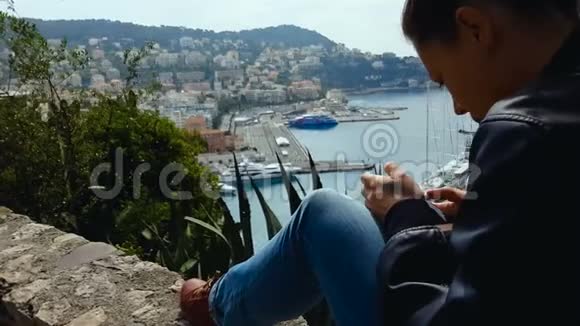 女子滚动智能手机坐在美丽的尼斯港自然城市景观视频的预览图