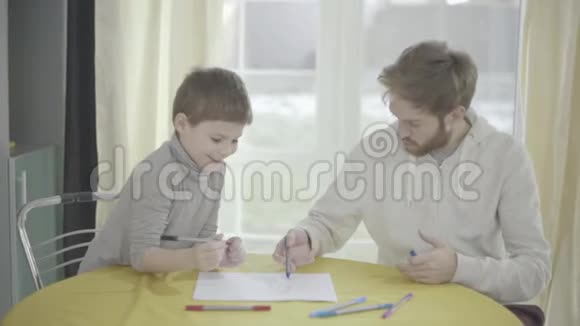 满脸胡须的父亲和他的小儿子坐在客厅的桌子上画着纸父子关系视频的预览图