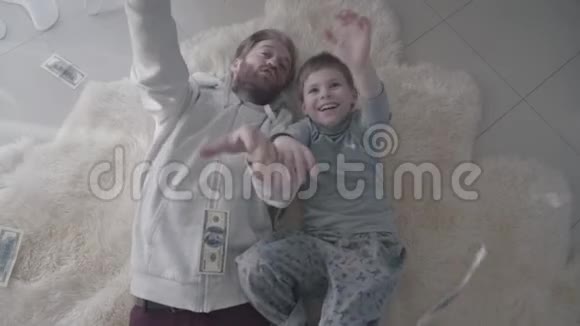 快乐的大胡子父亲和快乐的儿子躺在白色的毛皮地毯上抓着从上面掉下来的钱娱乐爸爸视频的预览图