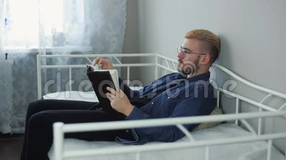 满意的年轻迷人的男人带着眼镜和黄色的头发在家躺在床上看书视频的预览图