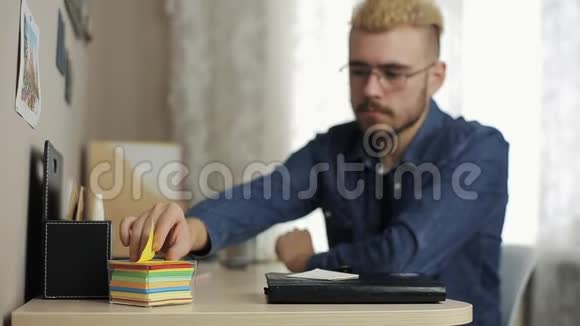 年轻的男学生发型时尚戴着眼镜用笔记本电脑和日记在桌子上的贴纸上做笔记视频的预览图