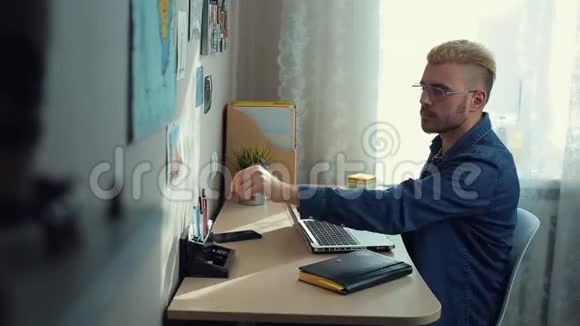 性格开朗的年轻男生带着眼镜和黄毛在家工作办公桌熟练的家庭自由职业者做笔记视频的预览图