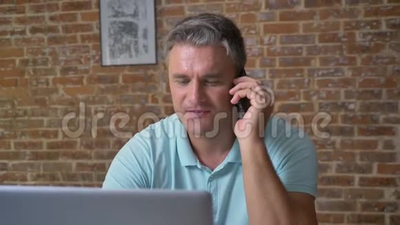 精力充沛的白种人成年男性在办公室里坐着用电话和电脑说话棕色的砖块视频的预览图