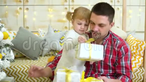 可爱的小女孩和年轻的父亲在家附近的圣诞树旁打开礼物盒视频的预览图