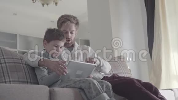 父亲和孩子用数字平板电脑坐在大客厅的沙发上爸爸教他的孩子快乐的爸爸和爸爸视频的预览图