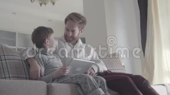 胡子爸爸和小儿子坐在窗户附近的沙发上在大客厅里玩平板电脑爸爸视频的预览图