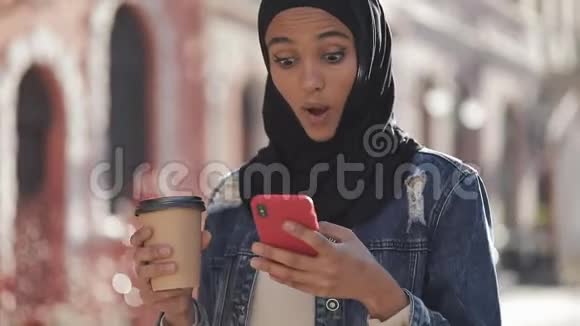 年轻美丽的穆斯林妇女使用智能手机行走在旧城市的背景她在智能手机上有好消息视频的预览图