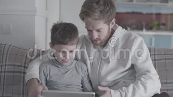 父亲和小儿子坐在大客厅的沙发上玩平板电脑爸爸教他的孩子父子关系视频的预览图