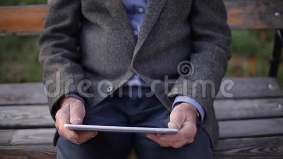 英俊的白发老人长着美丽的胡须坐在长凳上用平板电脑在网上滚动视频的预览图