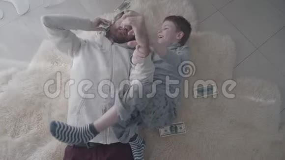 快乐的父亲和儿子在白色的毛皮地毯上玩钱玩得很开心娱乐爸爸和孩子父亲视频的预览图