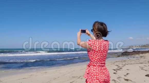 年轻迷人的女人拿着相机电话为矗立在塞浦路斯海滩上的风暴海浪拍照视频的预览图