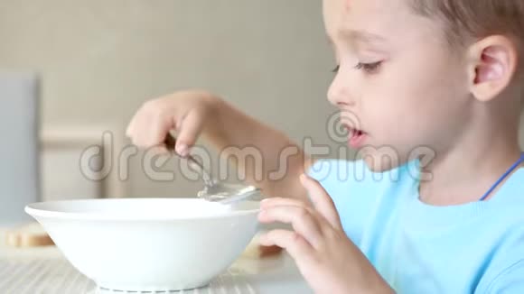 孩子坐在厨房的桌子旁用白色盘子里的勺子吃汤视频的预览图