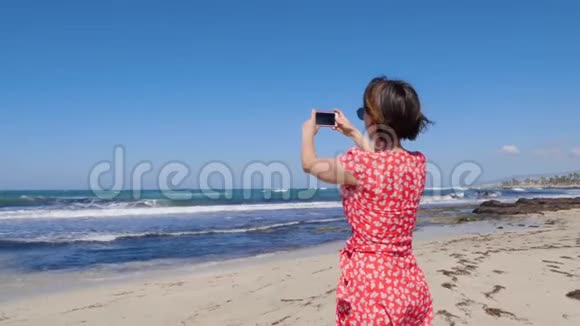 年轻迷人的女人拿着相机电话为矗立在塞浦路斯海滩上的风暴海浪拍照慢动作视频的预览图