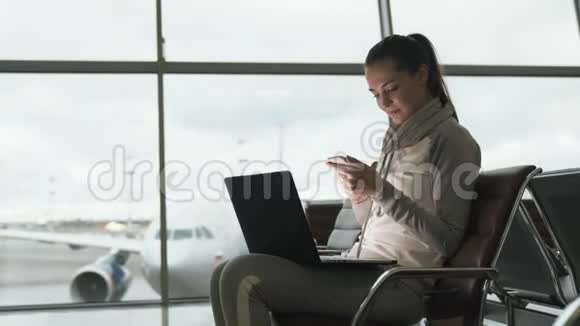 在候机室等待登机时漂亮女孩的侧视使用电话和笔记本电脑在机场工作视频的预览图