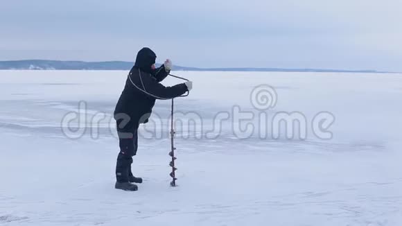 穿着深色衣服的渔夫用冰钻在冰冻的河流上挖洞视频的预览图