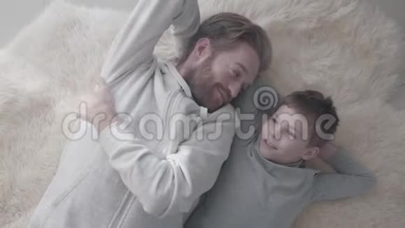 满脸胡须的父亲和他的小儿子躺在白色的毛皮地毯上进行了有趣的谈话爸爸说视频的预览图