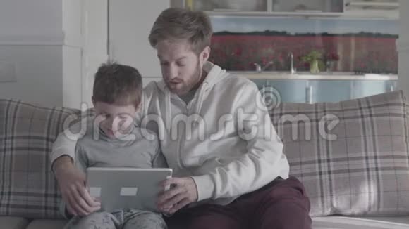 大客厅里长胡子的父亲和孩子用数码平板电脑坐在沙发上爸爸教他的儿子快乐视频的预览图