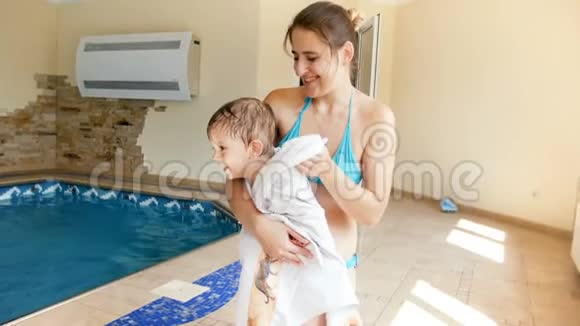 4k镜头微笑的年轻妈妈在游泳池里下课后用大毛巾擦干和温暖她蹒跚学步的儿子视频的预览图