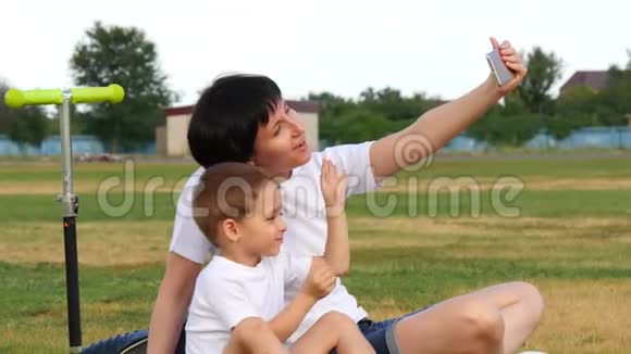 快乐妈妈和宝宝正坐在公园的绿草上用手机自拍视频的预览图