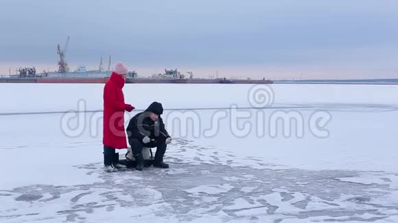 渔夫坐在冰冻的河流上在冬天的钓竿的帮助下钓鱼旁边是一位漂亮的年轻女子看着渔夫视频的预览图