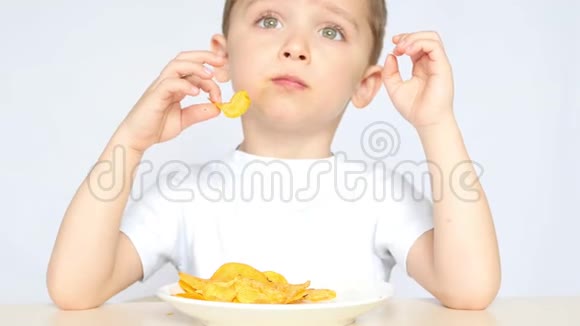 一个高兴又高兴的孩子吃着薯片坐在桌子旁的白色背景上特写视频的预览图