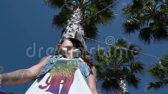 夏天时尚穿着时髦的年轻女士从下移到高大的树上在风中摇摆视频的预览图