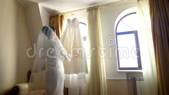 靠近窗户在房间里白色的婚纱挂在窗户的屋檐上新娘一个穿着白色睡衣的漂亮女孩视频的预览图