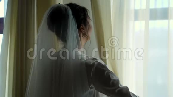 新娘的肖像美丽的女孩戴着面纱穿着白色的长袍推开窗帘望着窗外视频的预览图