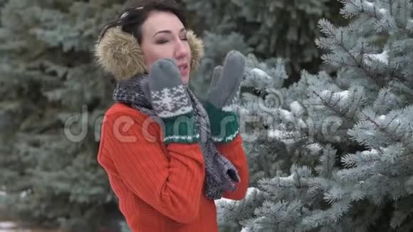 穿着毛皮手套和耳罩的女人摸着她的脸和脸颊冬季森林美丽的风景雪杉树视频的预览图
