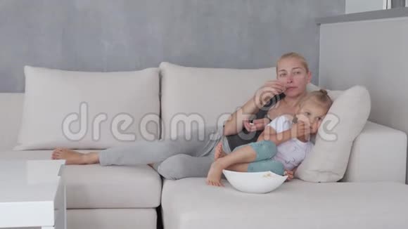 一个女人和一个小女孩坐在柔软的沙发上吃着爆米花看着电视屏幕视频的预览图