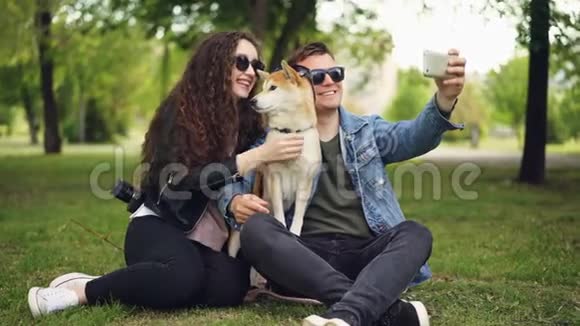 英俊的年轻人正在和他漂亮的妻子和可爱的狗自拍都戴着太阳镜他拿着智能手机视频的预览图