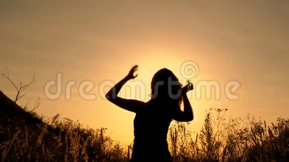 女孩跳起来用双手在田野里的黄色夕阳下在山峦之间的花丛中挥舞视频的预览图