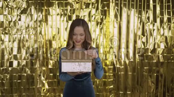 快乐兴奋的女孩打开一个盒子带着礼物金色的光从盒子里照在她的脸上一个金色背景的女人4K视频的预览图