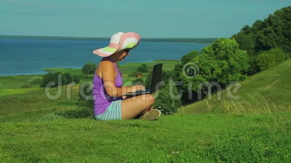 一个戴草帽的女人坐在俯瞰湖的山上关上笔记本电脑的盖子视频的预览图