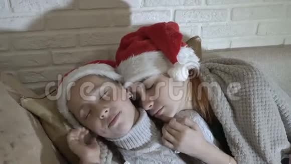 孩子们两个小女孩戴着圣诞老人的红帽子睡在沙发上盖着毯子视频的预览图