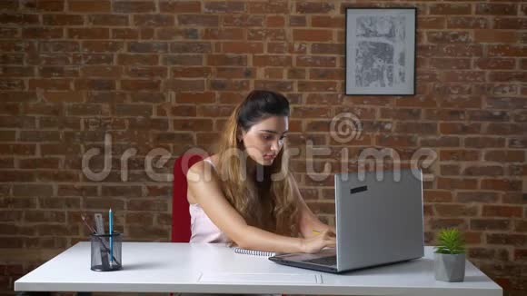 可爱的白种人长卷发女孩坐在桌面上从她的笔记本电脑上写下笔记红色的办公室视频的预览图