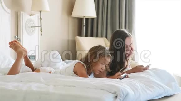 年轻的母亲和小可爱的女儿正在使用小工具躺在白色的床上视频的预览图
