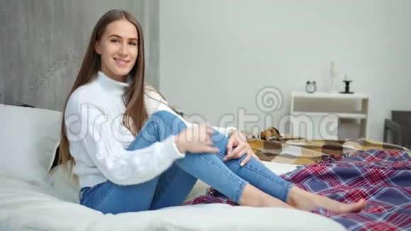 年轻漂亮的光着脚的年轻女子在卧室里坐在床上享受周末视频的预览图