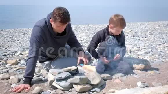 爸爸和儿子在海边的手工石头上点燃篝火中的煤视频的预览图