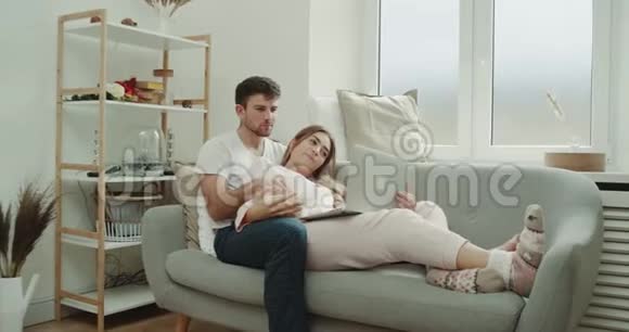 懒洋洋的一天一对年轻夫妇在沙发上吃爆米花穿着睡衣看着笔记本视频的预览图