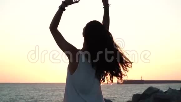 迷人的女孩留着黑色长发穿着一件透明的长袍在海滩上日落时摆姿势视频的预览图