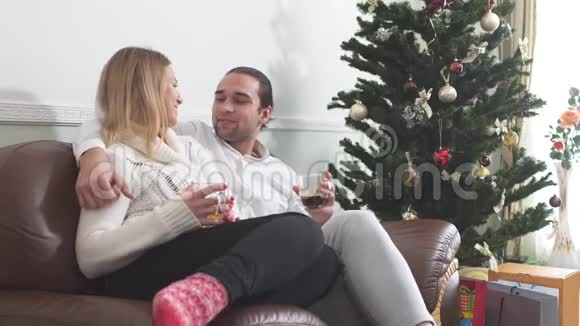 年轻人抱着他的女朋友坐在圣诞树旁边的沙发上喝咖啡恋爱关系视频的预览图