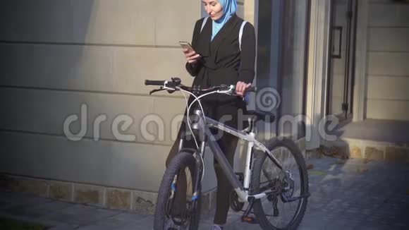 年轻的穆斯林妇女骑着自行车站在城市里用电话慢悠悠视频的预览图