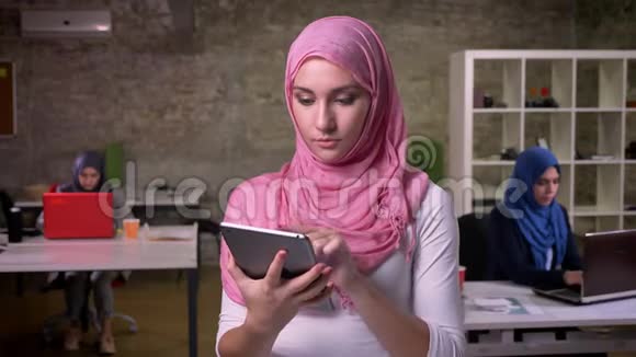 穿着粉红色头巾的漂亮微笑的女人站在砖制的办公室里坐着工作在平板电脑上显示着蓝色的屏幕视频的预览图