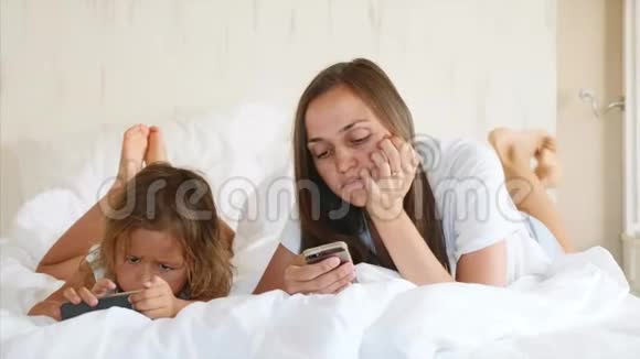 年轻的母亲和小可爱的女儿正在使用小工具躺在白色的床上视频的预览图