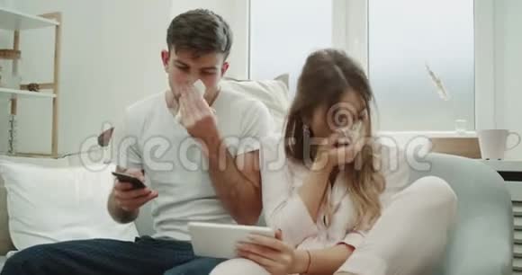 现代设计的年轻夫妇穿着睡衣躺在沙发上感觉不好患了流感流鼻涕用纸巾点了点什么东西视频的预览图